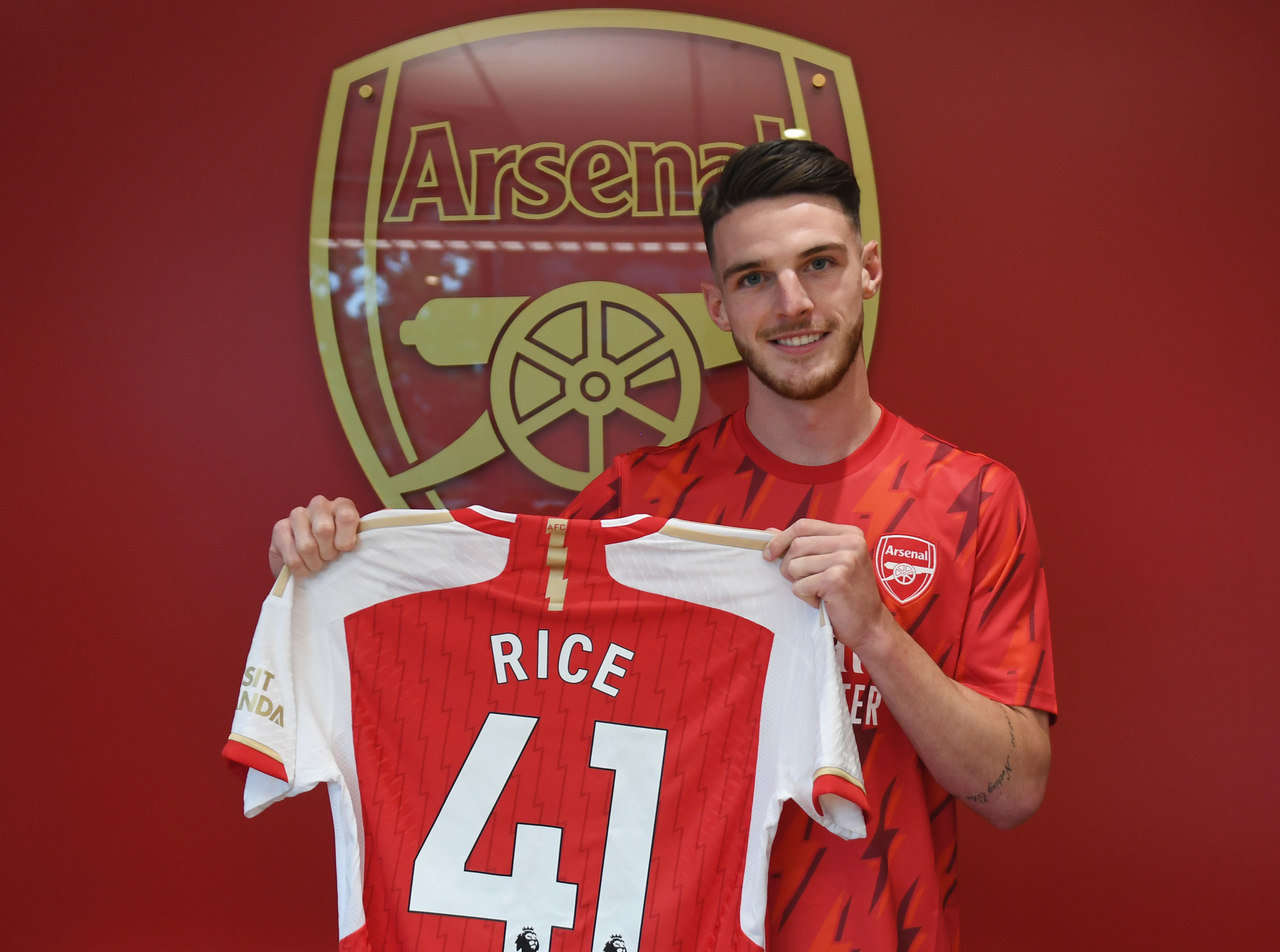  Declan Rice is pictured holding a jersey with his name and the number 41 on it, while standing in front of the Arsenal logo.