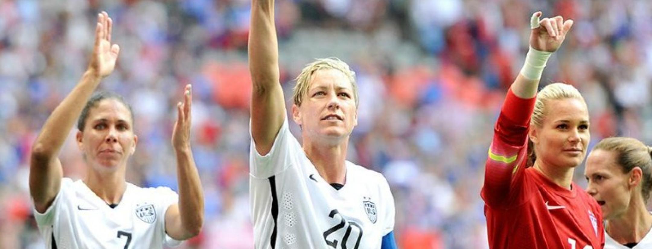 Abby Wambach and memebrs of the USWNT are shown with their arms raised in thumbs up.