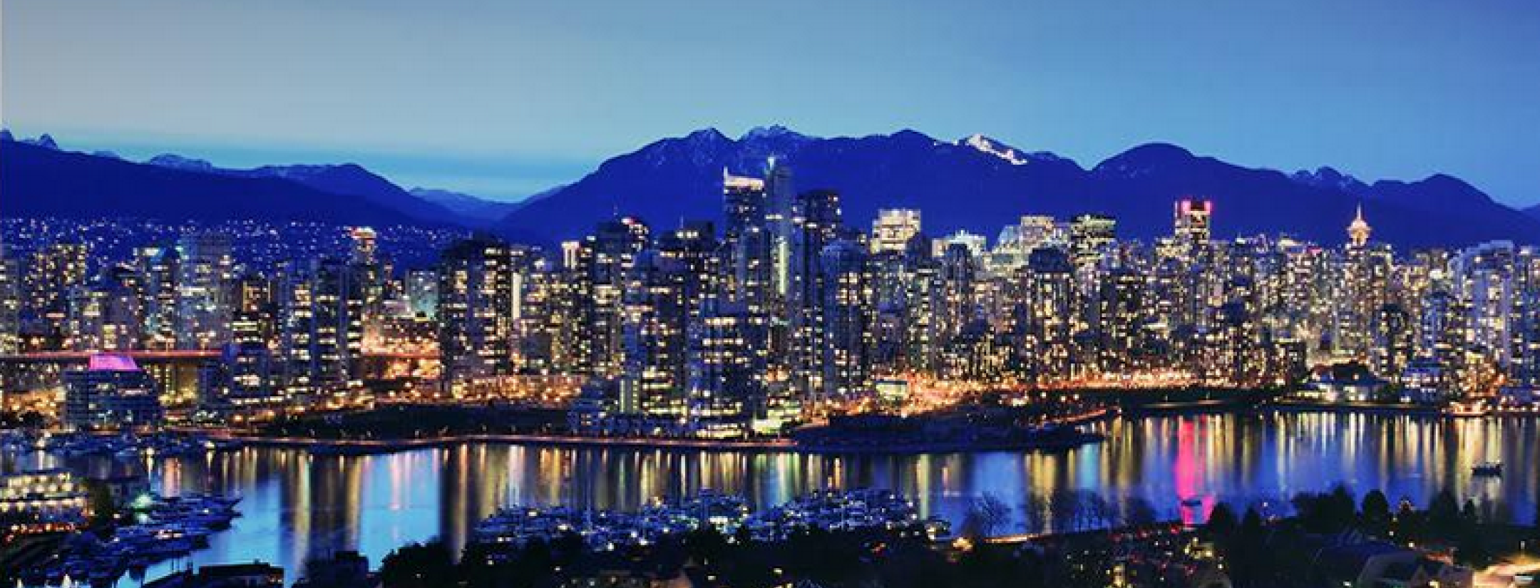 A night time view of beautiful Vancouver. The skyline is illuminated by its skyscapers countless lights infront of dark blue night sky and purple mountains. 